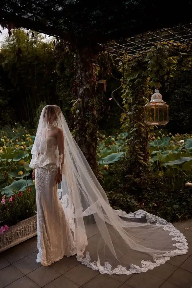 A bride in a flowing wedding gown and veil stands in a garden filled with greenery and lotus flowers, with a lantern hanging nearby.