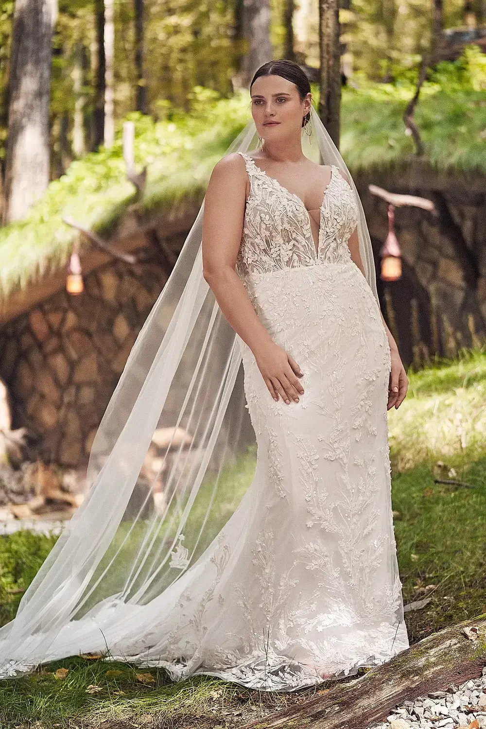 A woman in a detailed white wedding dress with a plunging neckline and a long veil, standing in a natural setting with trees and greenery in the background.