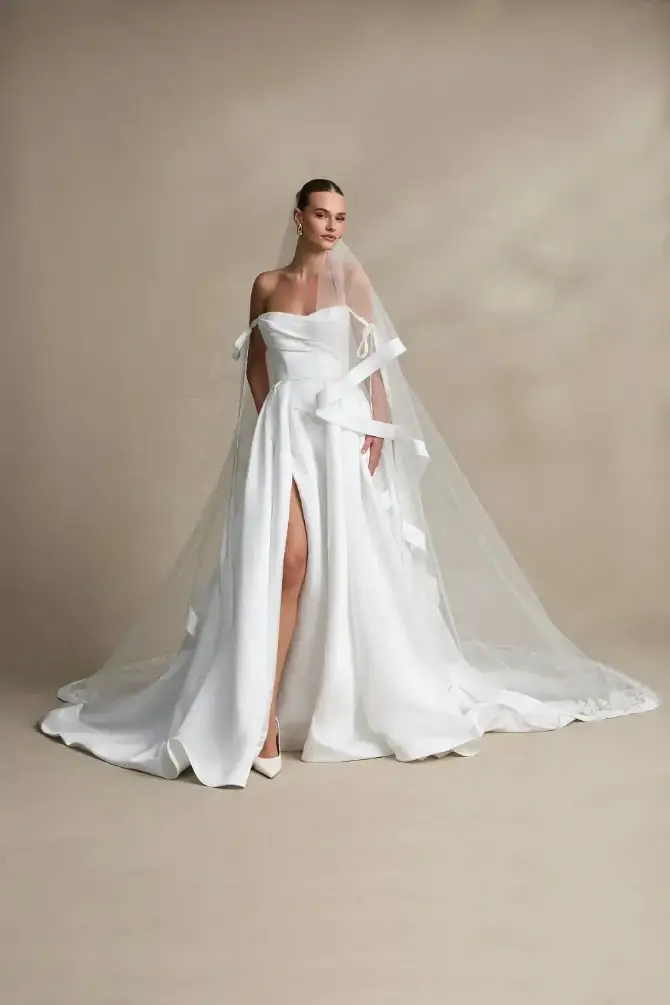 Elegant bride in a strapless white wedding gown with a front slit, wearing a long veil. She stands on a neutral backdrop, exuding grace and sophistication.