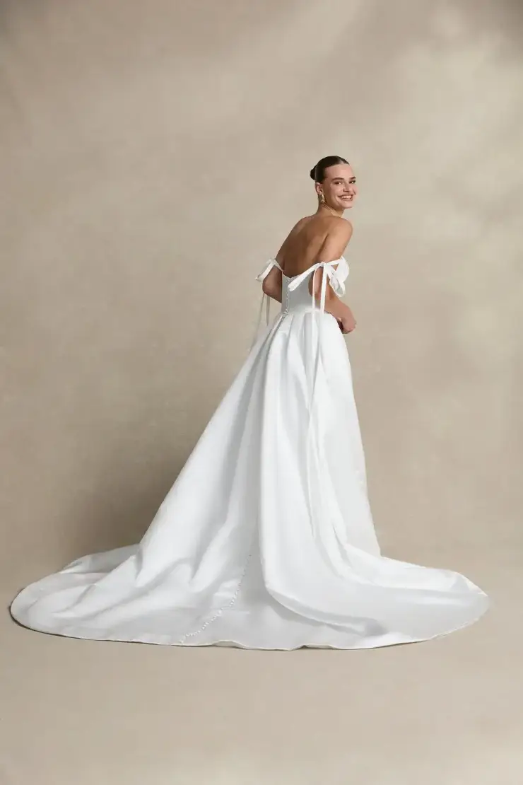 A woman in an elegant off-shoulder white wedding dress smiles while looking back. The dress has a long train and delicate bows, set against a neutral background.