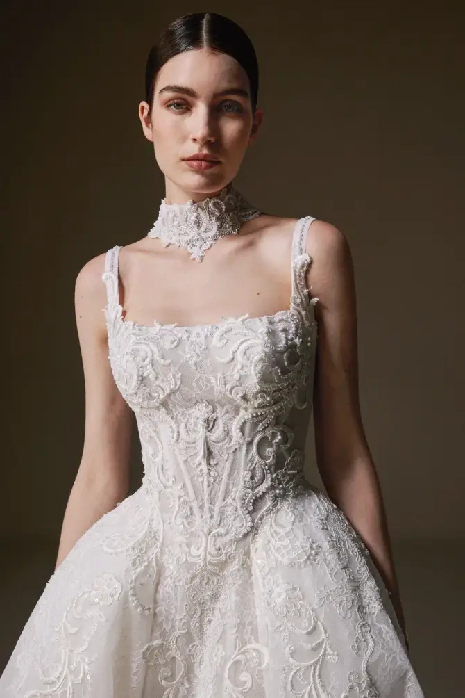 Woman wearing an ornate, white lace wedding dress with intricate embroidery and a matching lace choker. She stands against a neutral background, exuding an elegant and serene demeanor.