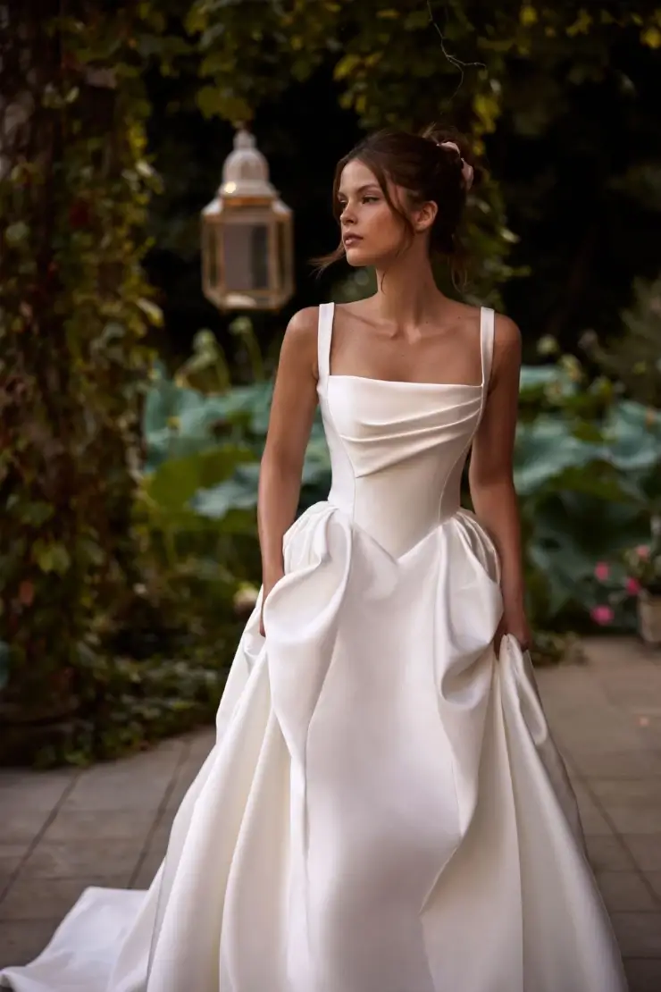 A woman in a white, elegant dress with unique detailing, standing outdoors surrounded by greenery.