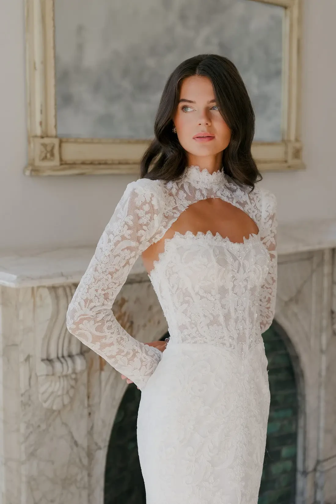A woman in a delicate lace wedding gown stands gracefully by a marble fireplace, exuding elegance and confidence, with soft lighting and neutral tones.