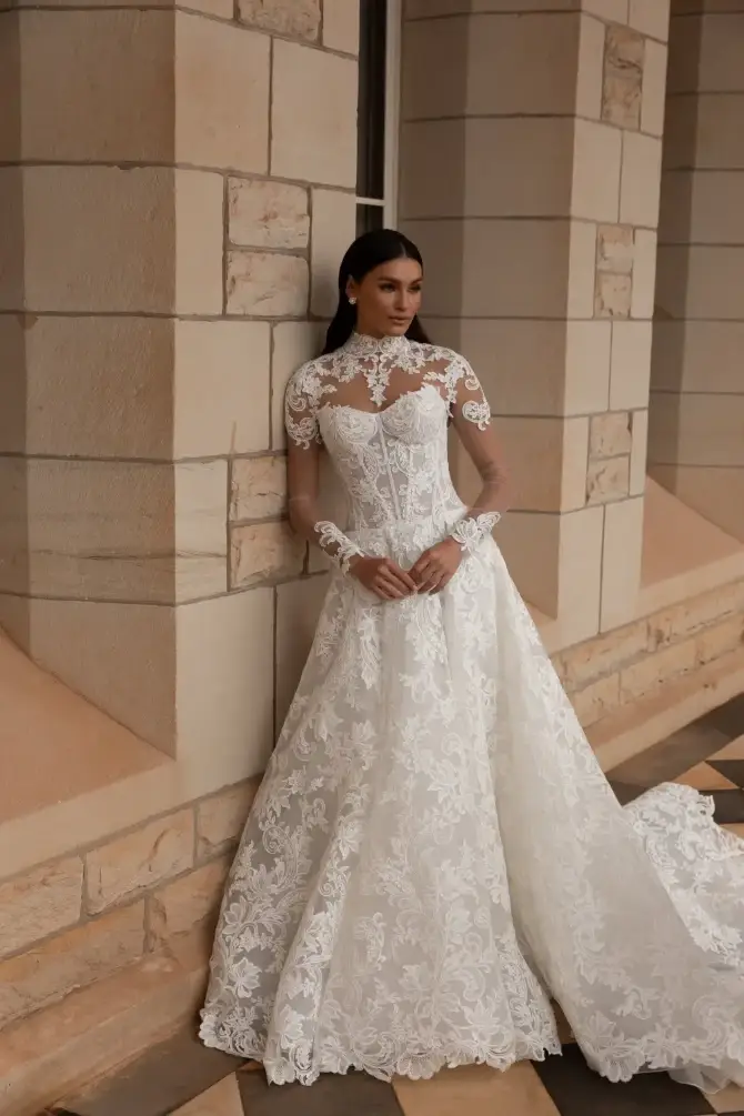 Model is wearing a strapless drop waist lace wedding gown with a corset bodice and removable illusion long sleeve lace high neckline bolero