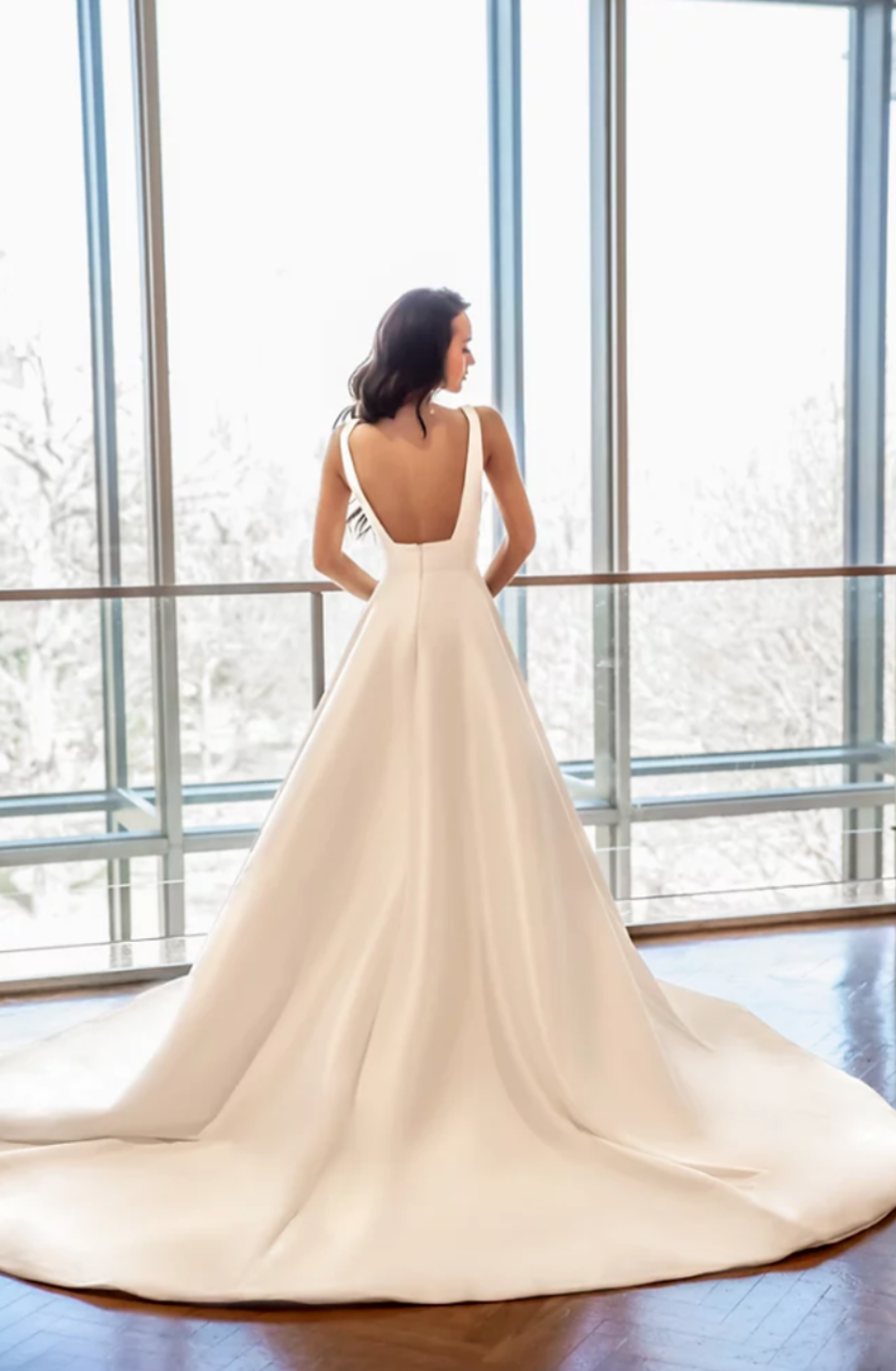 Model is wearing a dove satin gown featuring a flattering v-neck bodice, wide waistband, and illusion v-sides.