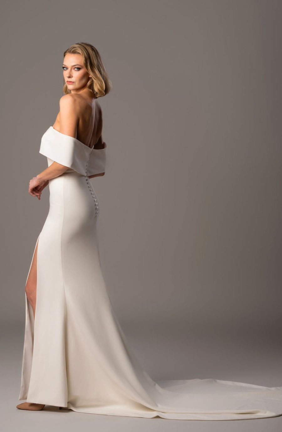 Model is wearing a fit-and-flare wedding dress with off-the-shoulder sleeves, a paneled bodice, high slit, and fabric-covered buttons trailing down the back.