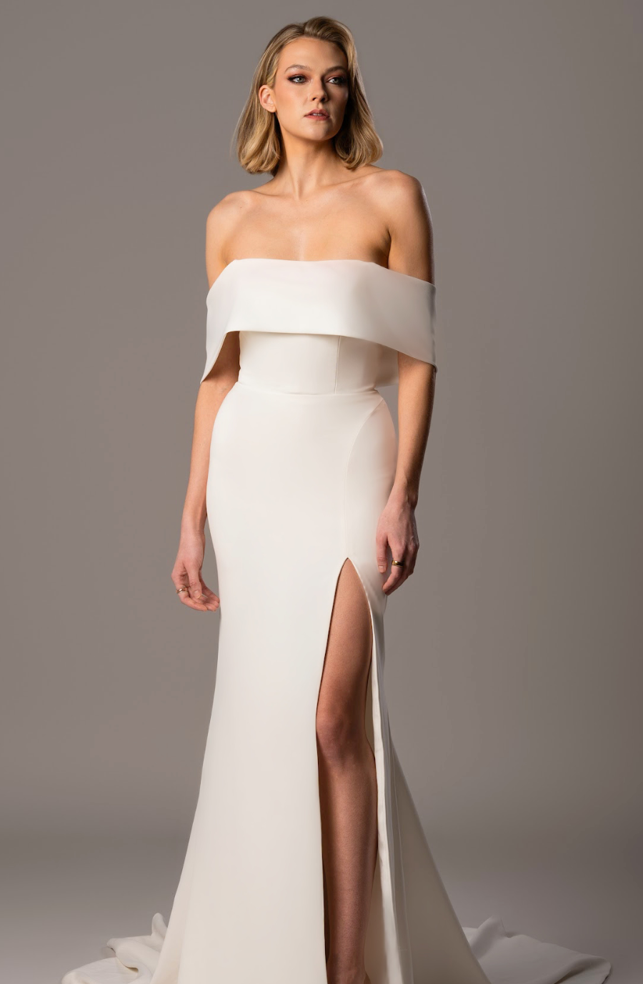 Model is wearing a fit-and-flare wedding dress with off-the-shoulder sleeves, a paneled bodice, high slit, and fabric-covered buttons trailing down the back.