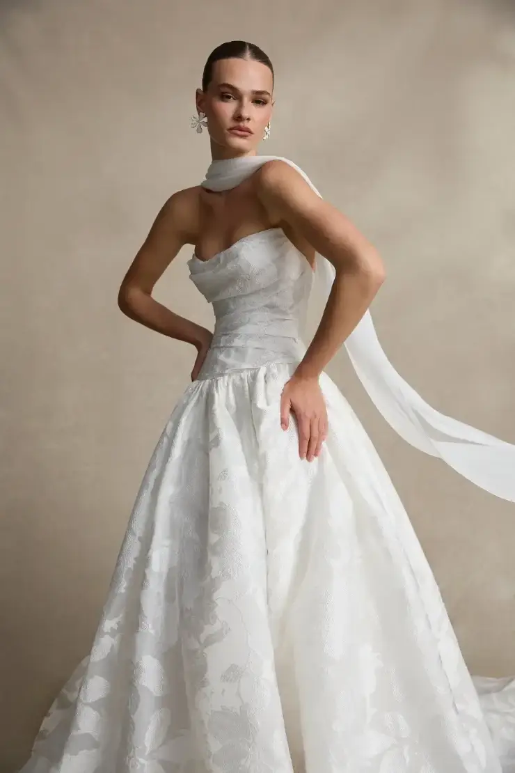 Model is wearing a strapless dropped waist wedding dress with a full jacquard skirt, draped bodice, and bridal scarf around the neck