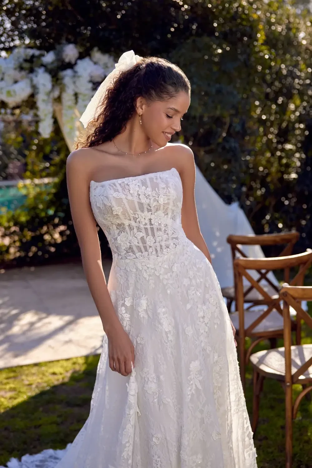 A woman in a strapless white wedding dress stands outdoors, smiling, with her hair styled in soft curls and a veil.