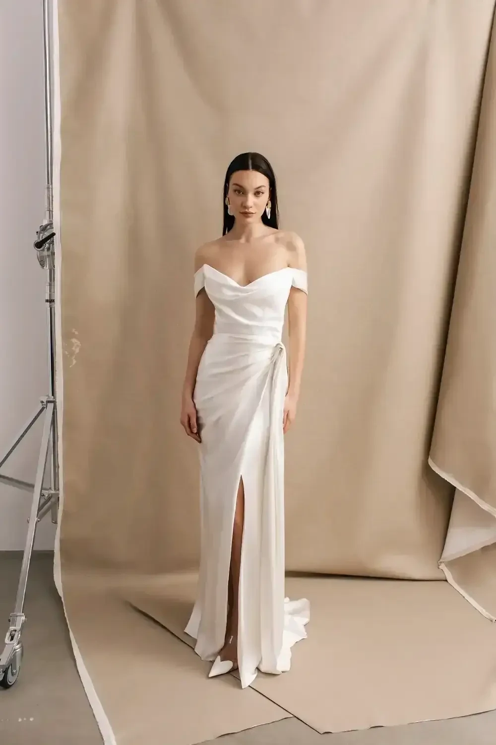 A woman in an elegant off-the-shoulder wedding dress, smiling softly while looking to the side, with long dark hair and wearing earrings, against a backdrop of soft, light curtains.