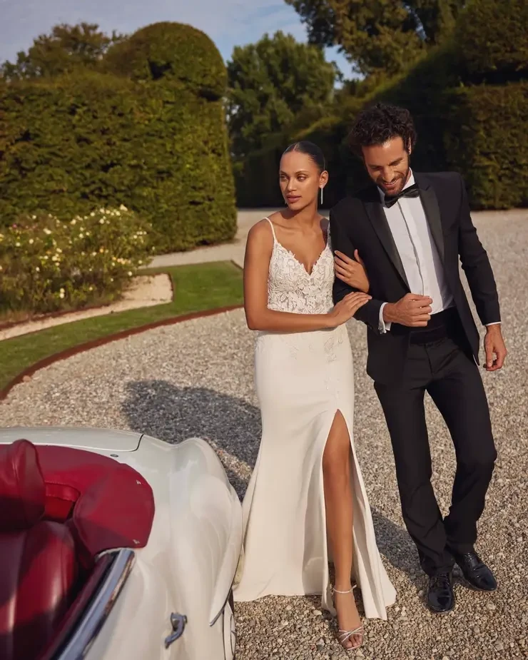 Model is wearing a figure-flattering fit and flare gown in stretch crepe with a sheer bodice, sequined lace appliqués, scoop back, and optional slit.