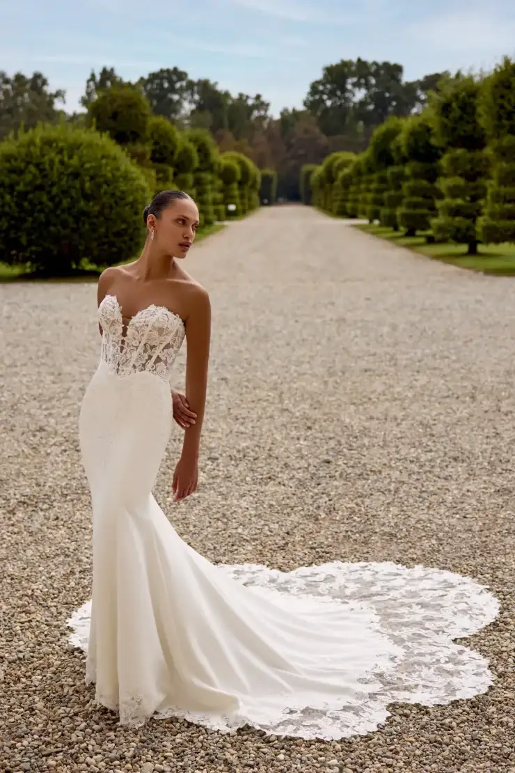 A model wearing a stunning bridal gown with a fitted silhouette and intricate lace detailing, posing elegantly in a scenic outdoor setting with manicured hedges and gravel pathways.