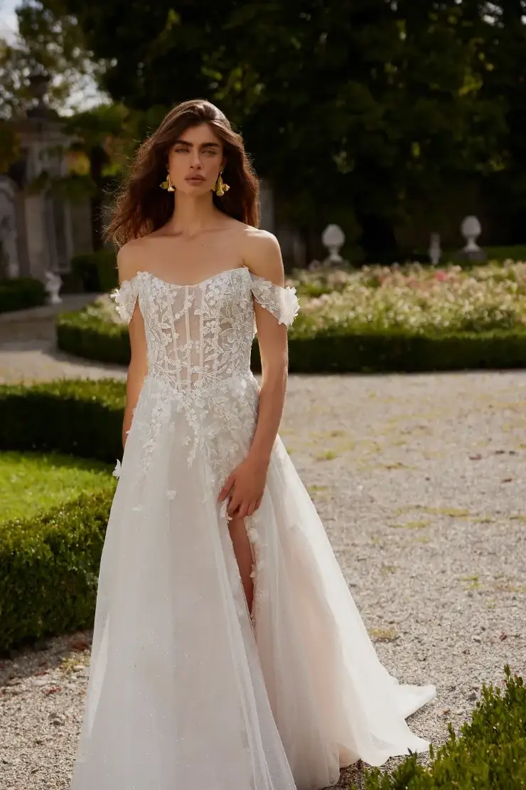 A model wearing an elegant off-the-shoulder wedding gown with intricate floral details and a high slit, standing in a beautifully landscaped garden.