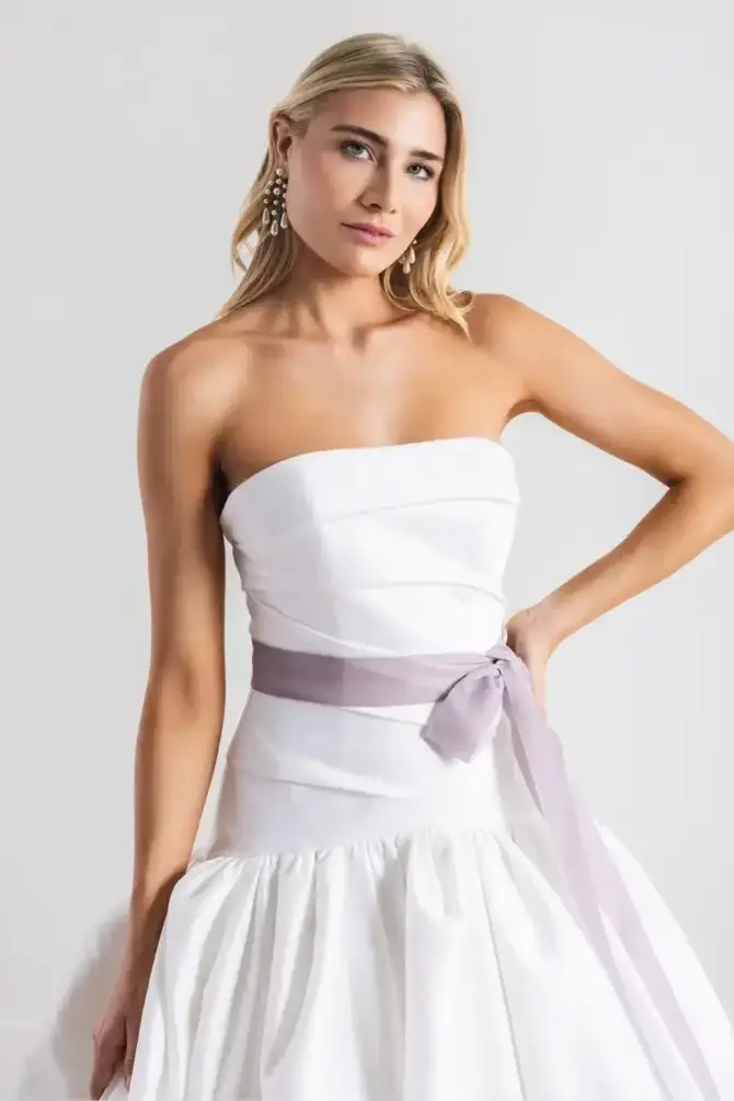 Model is wearing a strapless a-line dropped waist wedding dress with a light purple sash tied around the waist