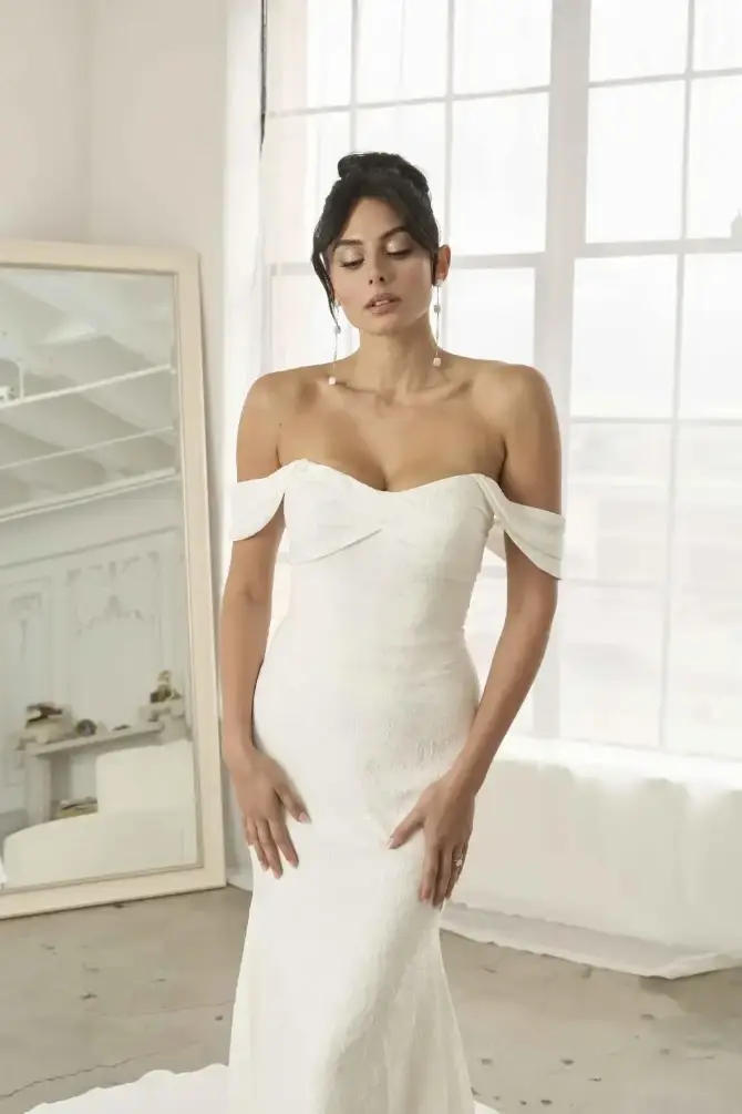 A woman in a strapless white dress stands gracefully in a well-lit room with large windows. She has her hands gently resting on her hips and appears to be deep in thought. A mirror is visible in the background.