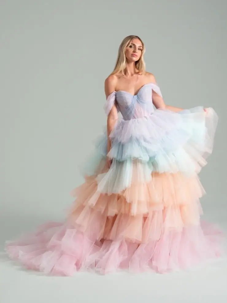 A woman in a strapless pastel gown stands against a plain background. The layered tulle dress features hues of pink, blue, and peach, creating an ethereal and elegant look.