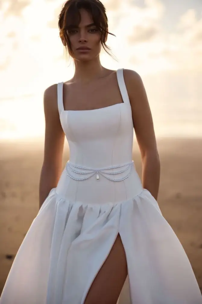 Model is wearing a sleeveless square neckline drop waist wedding dress with a thigh high slit and pearl belt around the waist